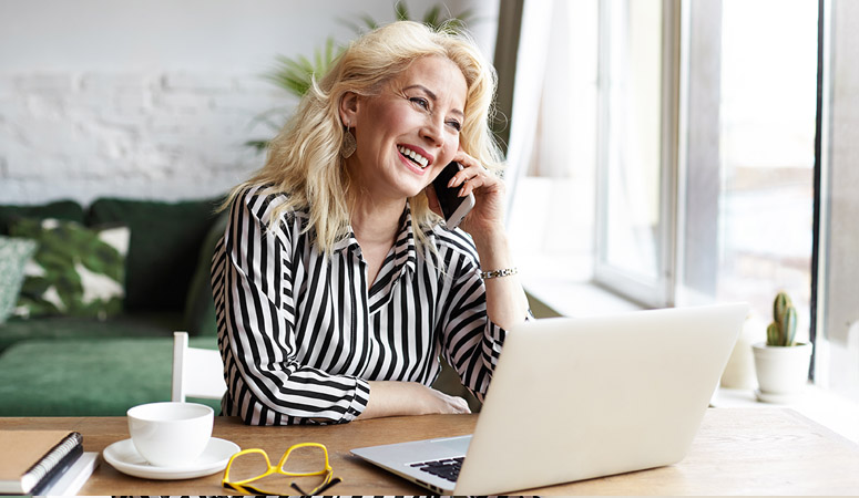 Attractive successful elderly businesswoman in striped blouse working in modern office, making phone call to potential client, having nice conversation, sitting at desk in front of open laptop